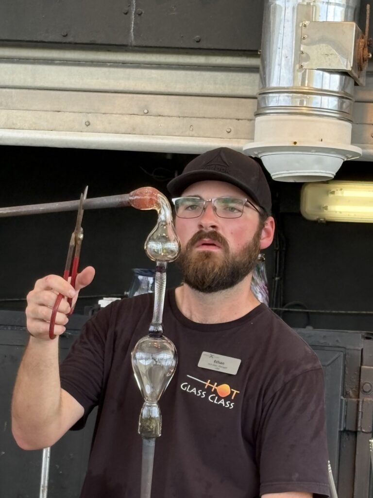 Glass artist Ethan Ball making a handblown glass goblet during a live glassblowing demonstration using traditional furnace techniques